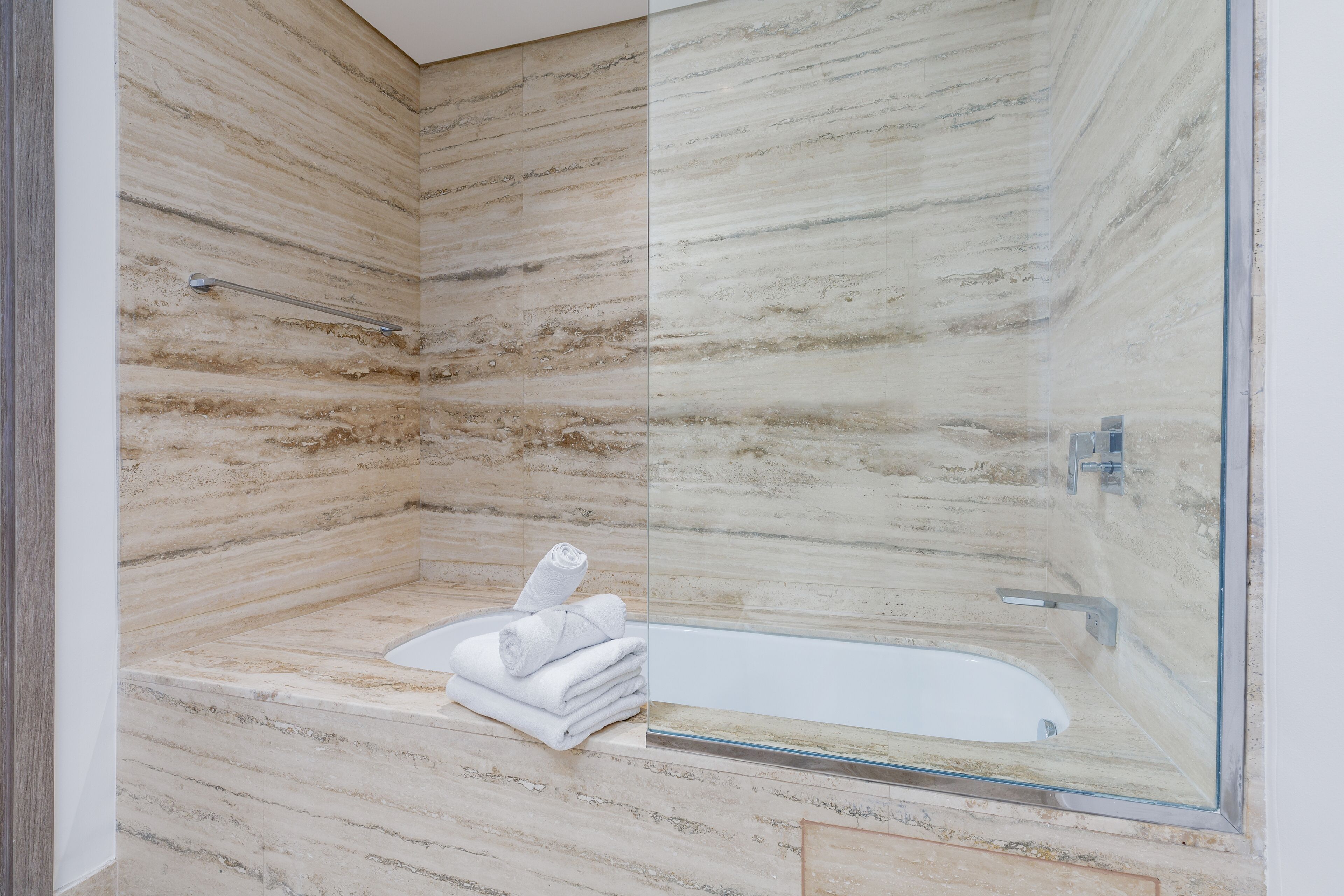 Un baño moderno con pared de mármol, cabina de ducha de vidrio y bañera con una pila de toallas.