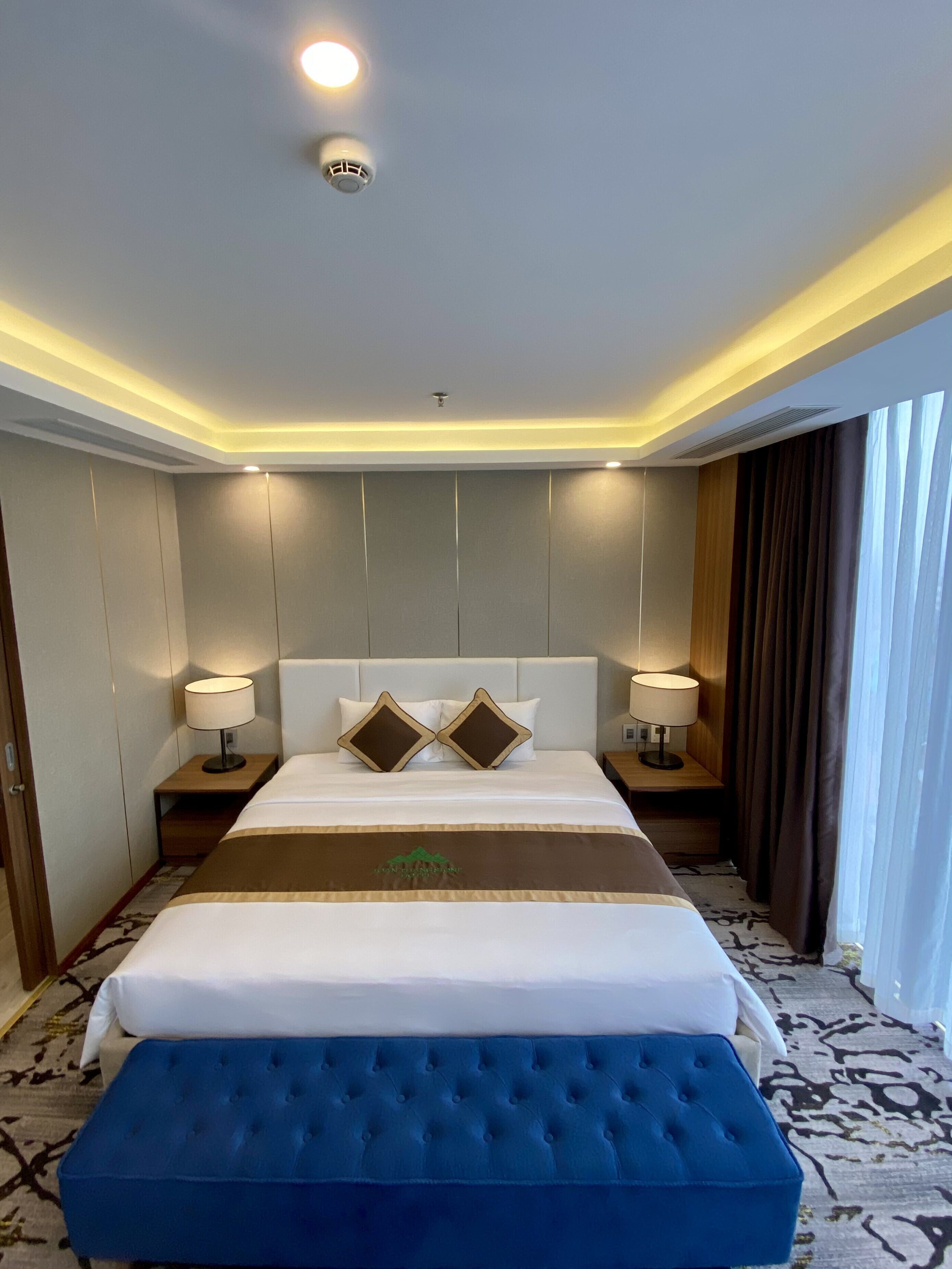 A hotel room with a large bed, two bedside tables with lamps, a ceiling light, and a window with curtains.