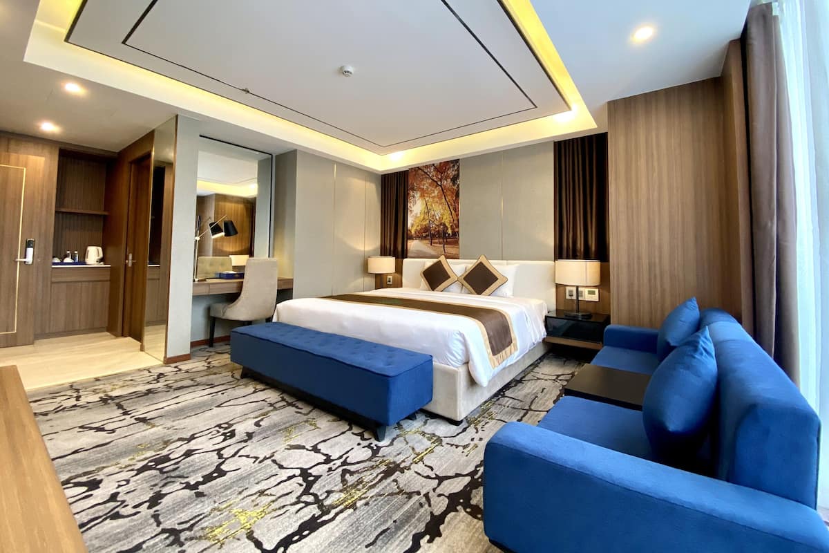 A modern hotel room with a large bed, a desk, a chair, a blue sofa, and a patterned carpet.
