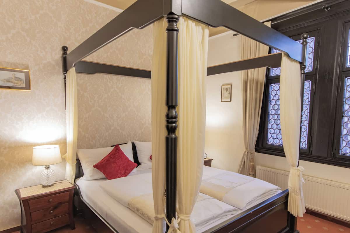 Una cama con dosel y cuatro columnas, una mesita de noche, un almohadón rojo y una lámpara.