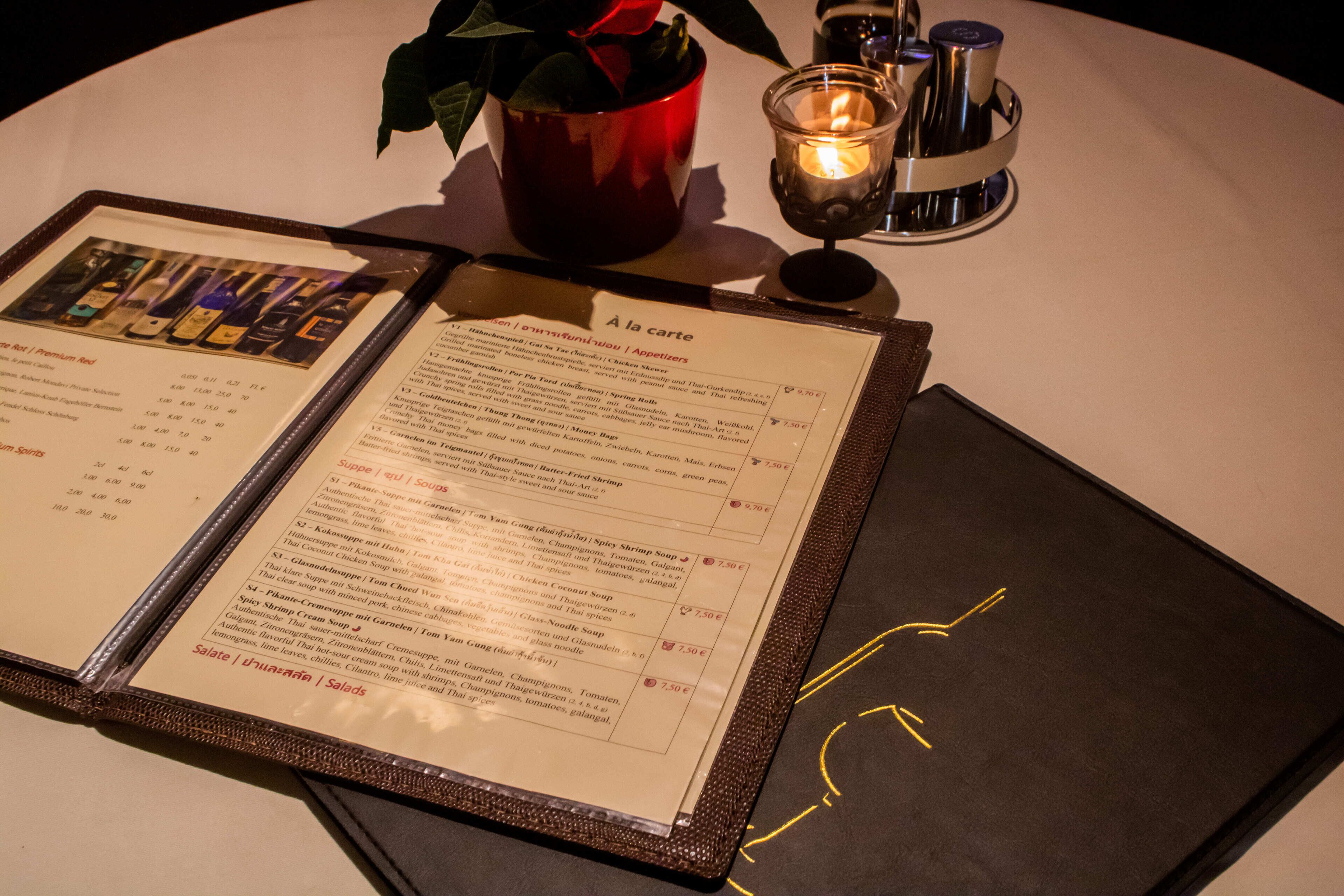 A menu with various dishes and prices, a candle, and a red cup with a rose on a table.