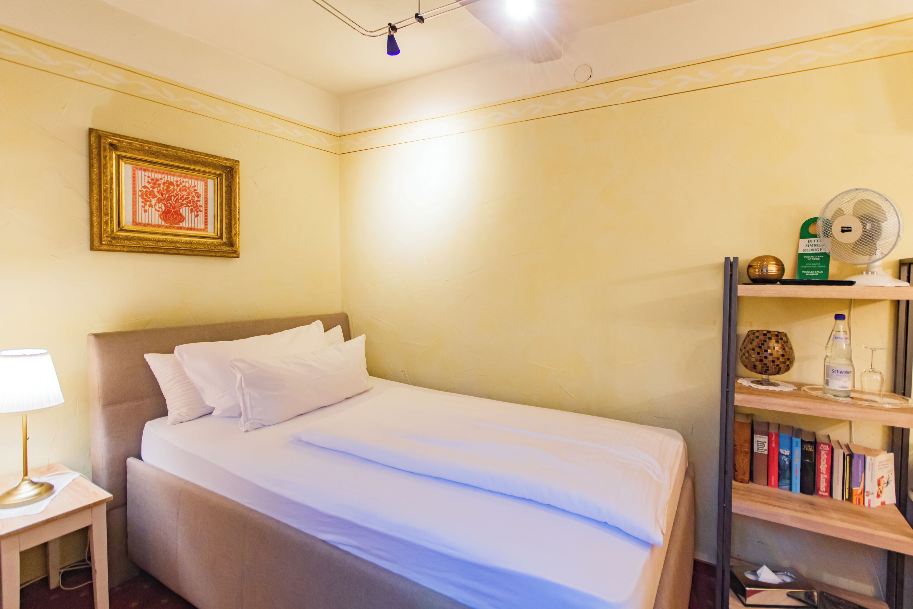 A neatly made bed with white linens, a bedside lamp, a framed artwork on the wall, a bookshelf with books and a fan, and a small decorative item on the shelf.