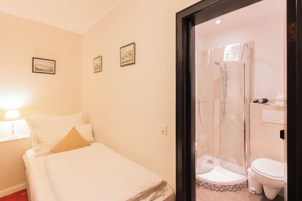 Habitación de hotel con cama, mesita de noche con lámpara, zona de ducha y retrete.