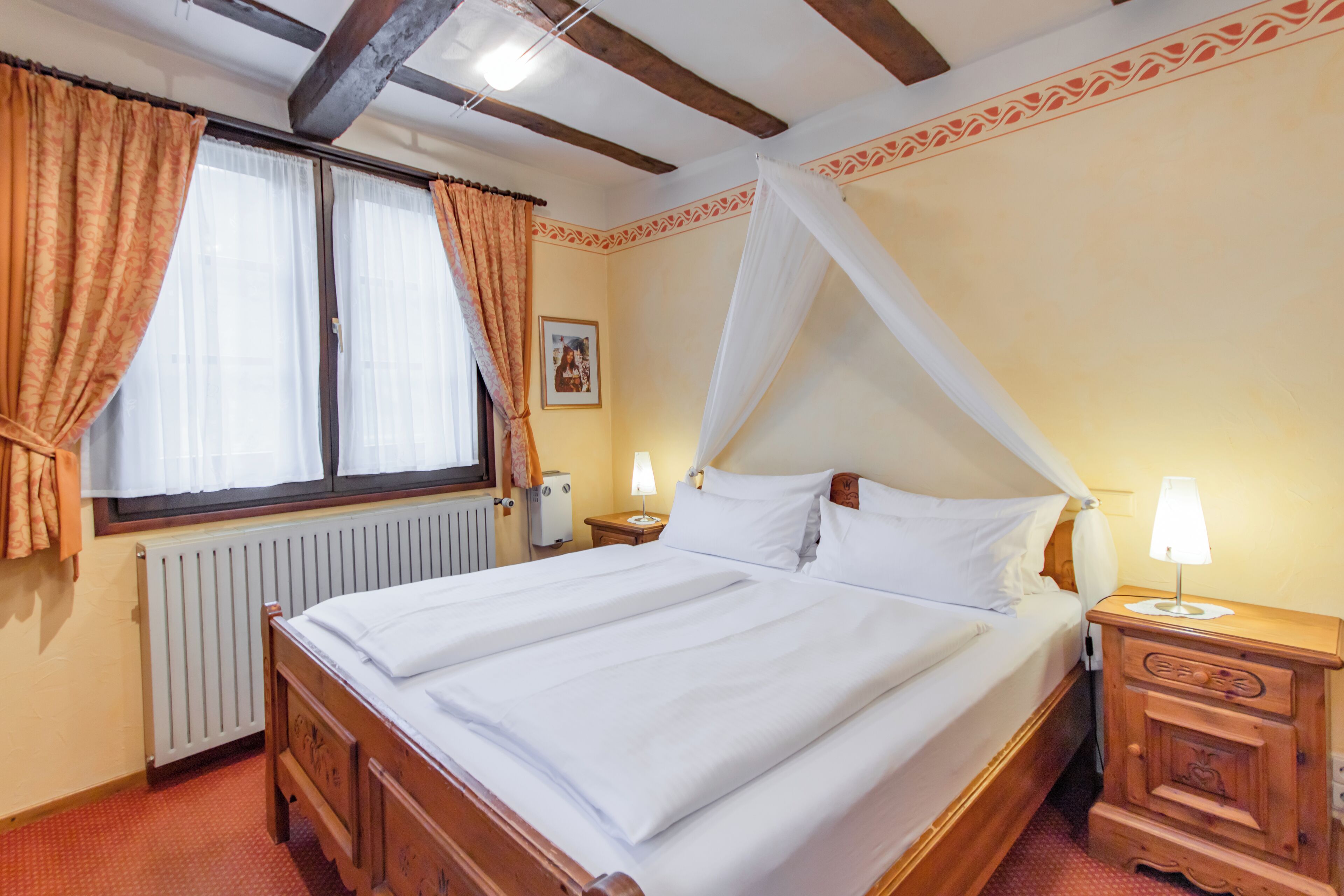 A double bed with white linens, a wooden headboard, a nightstand with a lamp, a radiator, and a window with curtains.