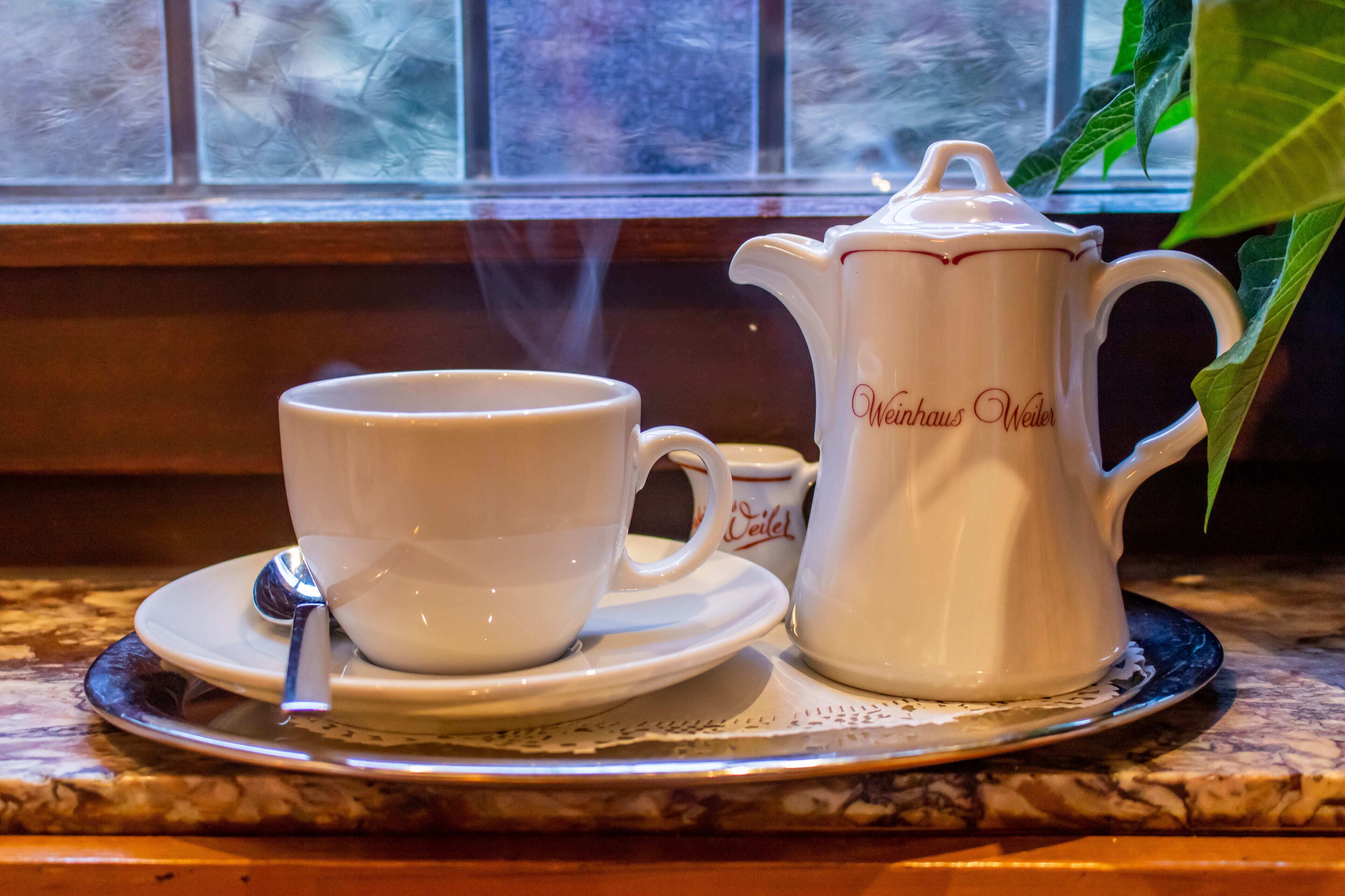 A white coffee pot and cup on a tray by a window with steam rising from the cup.