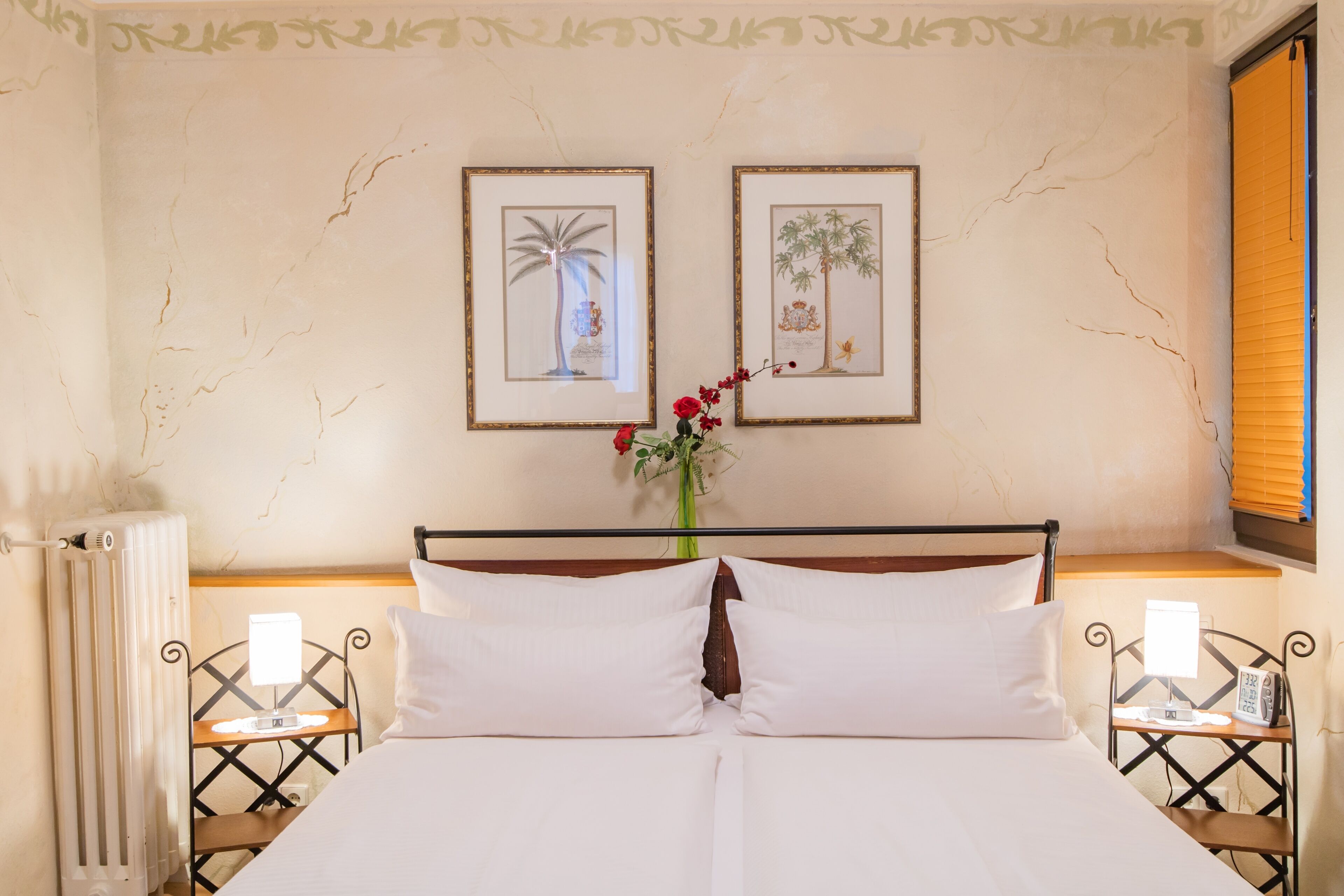 A neatly made bed with white linens, two framed pictures on the wall, a vase with red flowers, and a bedside table with a lamp and a phone.