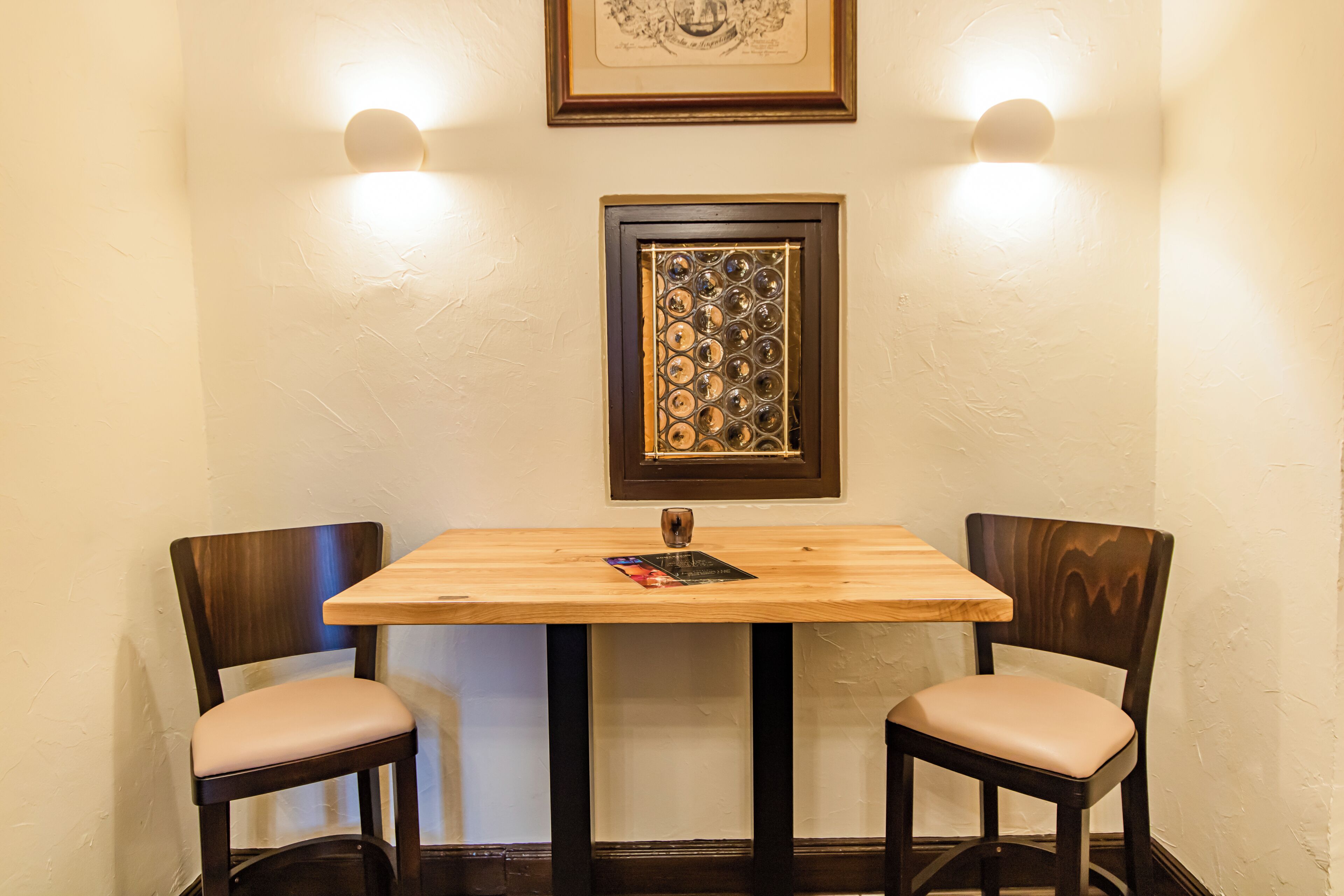 Un comedor con una mesa de madera y dos sillas, una obra de arte enmarcada sobre la mesa, y luces fijadas a la pared a cada lado.