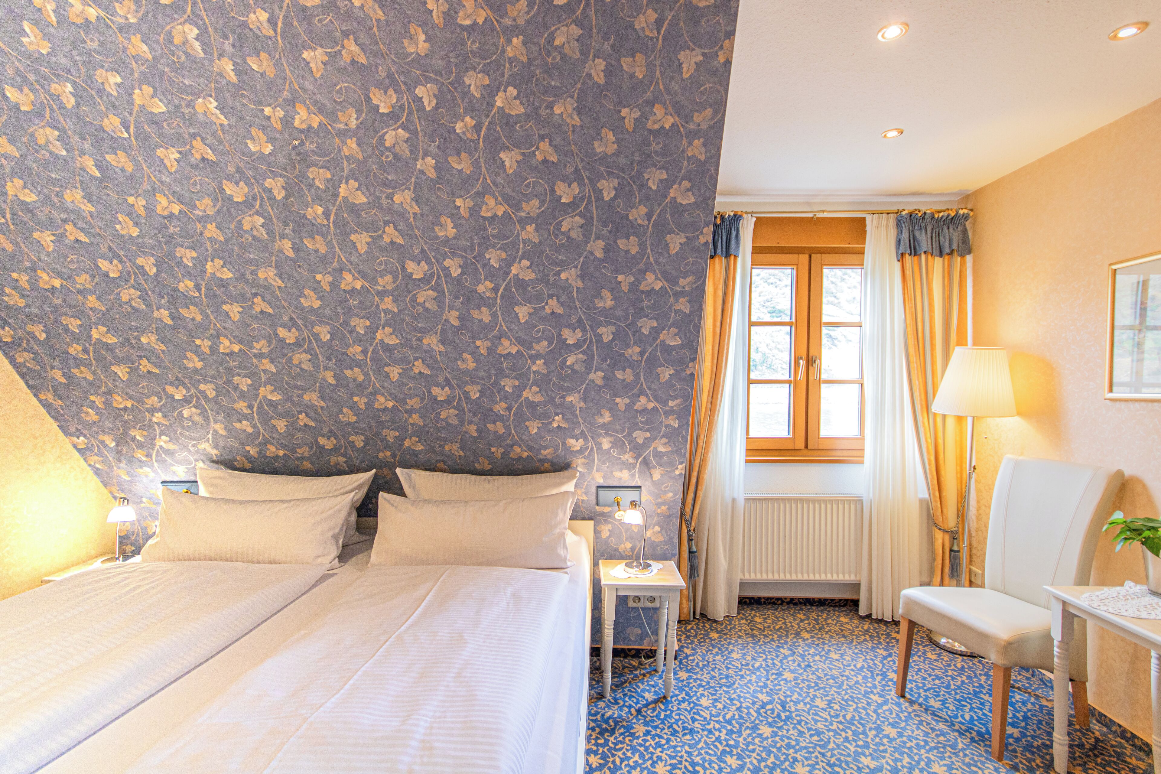 Ein Hotelzimmer mit einem großen Bett, einem Nachttisch, einem Sessel und einem Fenster mit Vorhängen.
