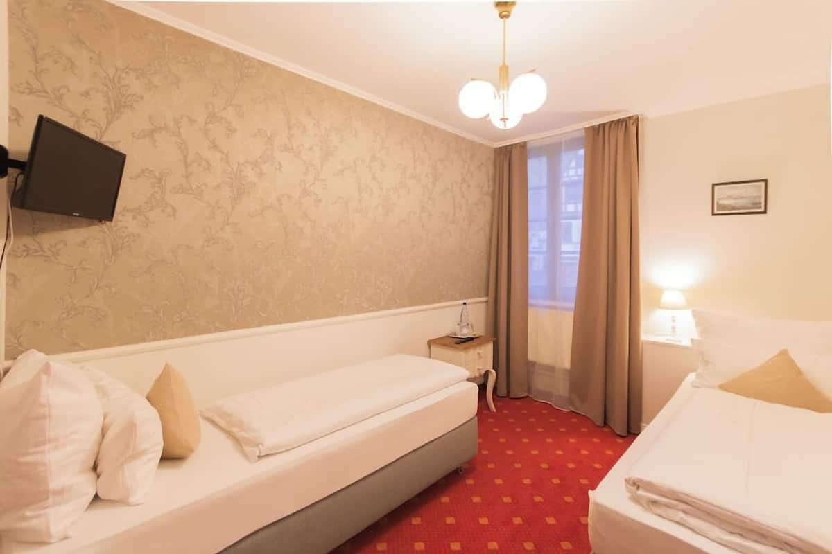 Habitación de hotel con dos camas, un televisor montado en la pared, una mesita de noche con lámpara y una ventana con cortinas.