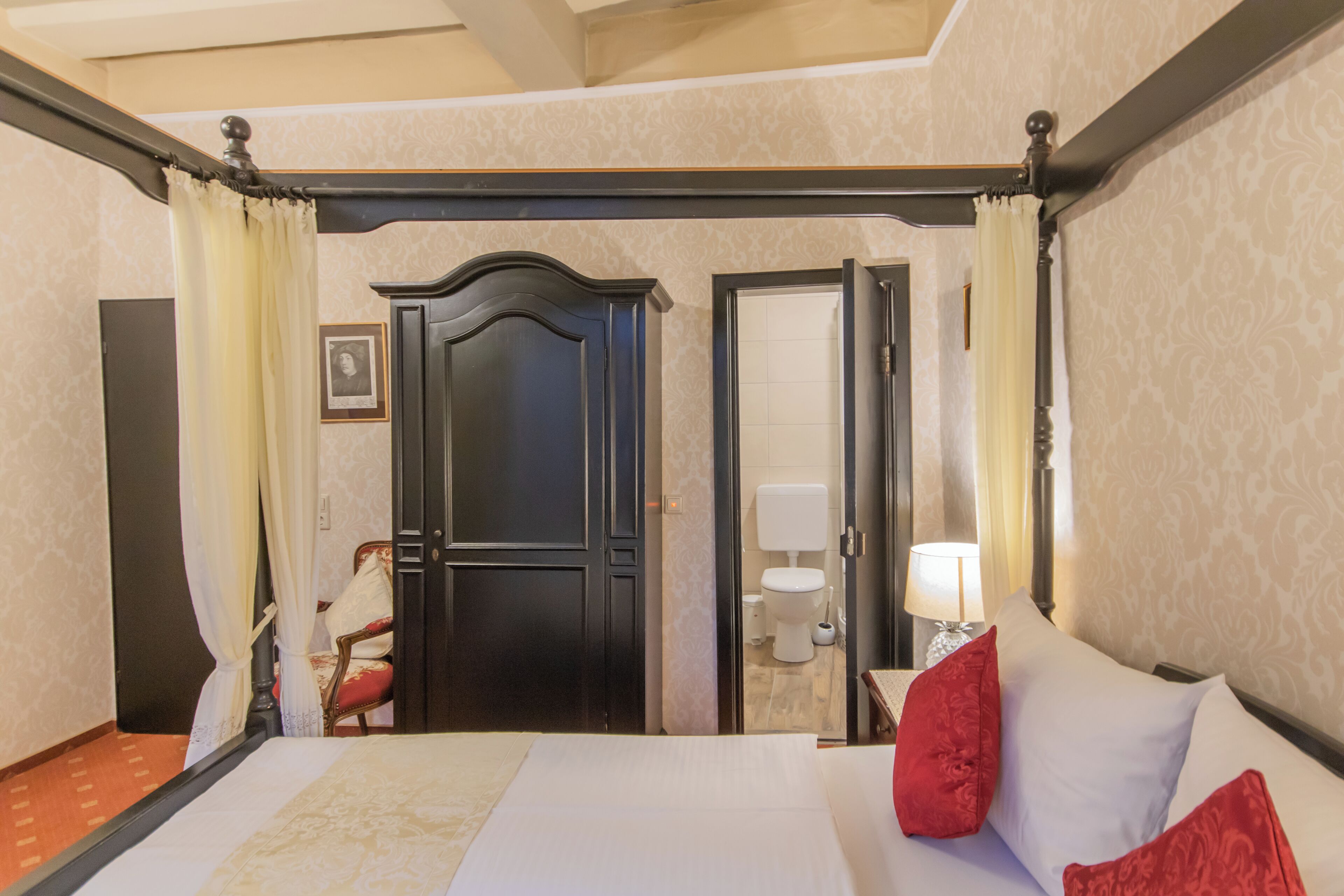 A hotel room with a four-poster bed, a wooden door, a bathroom with a toilet and sink, and a chair.
