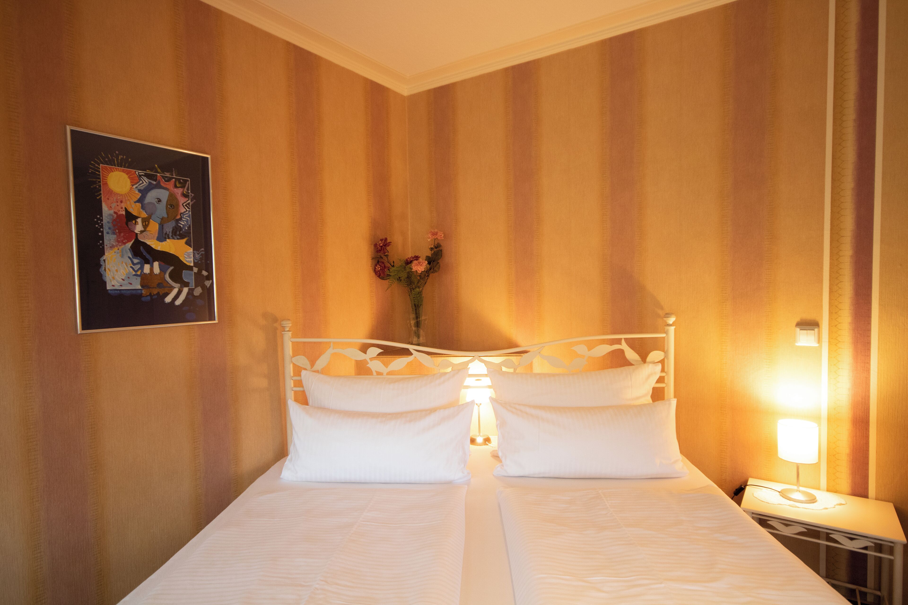 Una habitación de hotel con cama, mesita de noche, lámpara, un cuadro y un florero con flores.