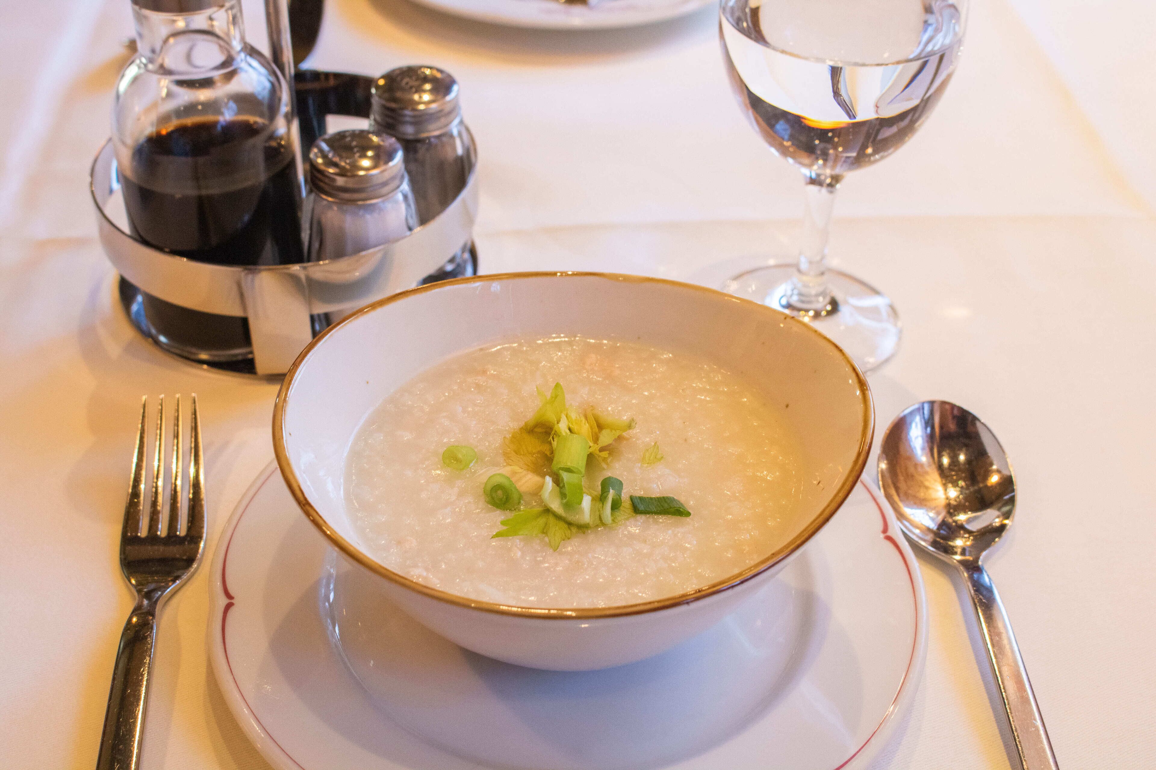 Un tazón de sopa adornado con cebollín, acompañado de un tenedor, cuchara y juego de salero y pimentero sobre una mesa.