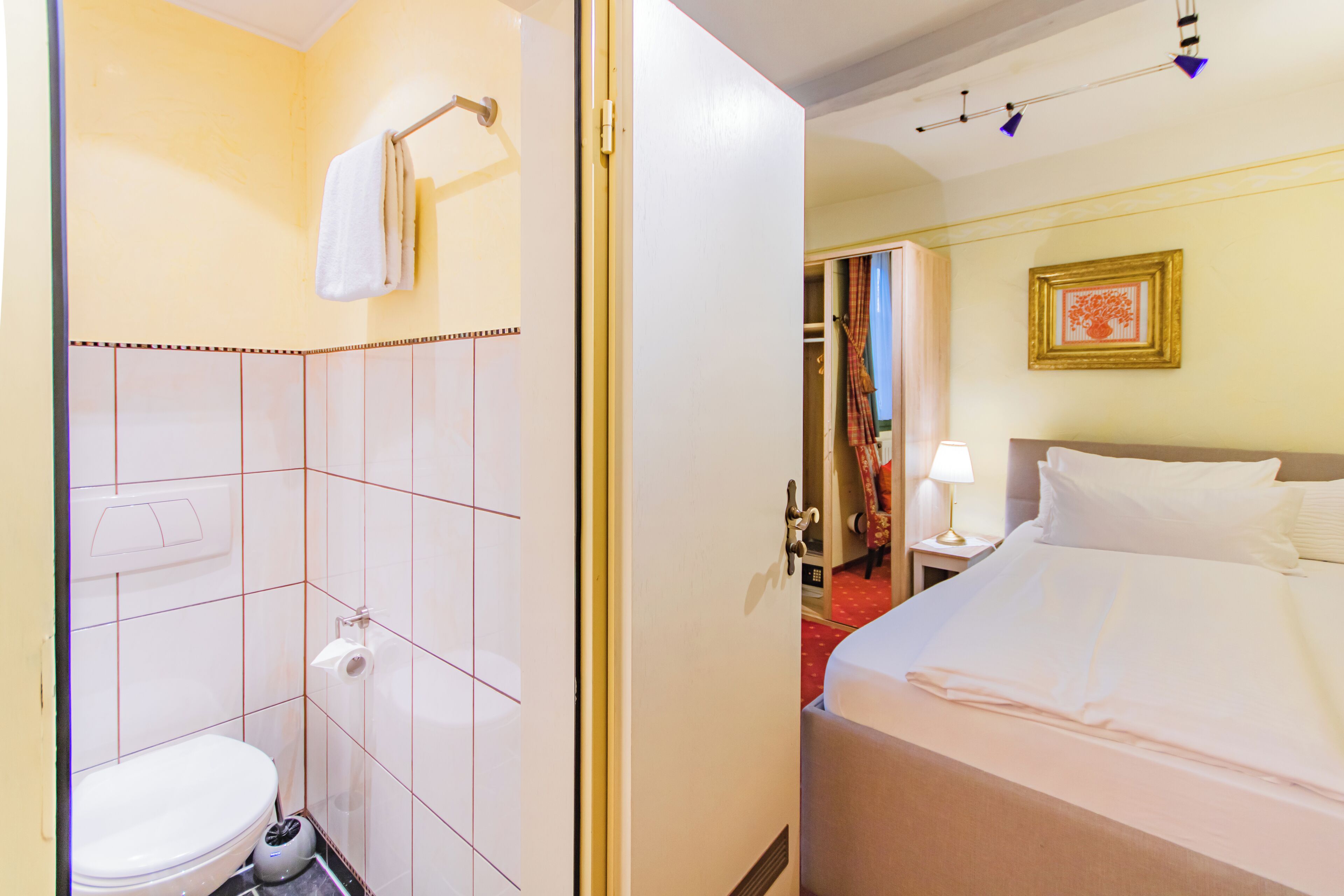 Quarto de hotel com sanita, uma cama, mesa de cabeceira com candeeiro e um quadro na parede.