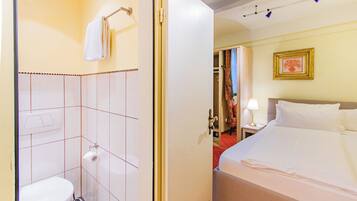 Quarto de hotel com sanita, uma cama, mesa de cabeceira com candeeiro e um quadro na parede.
