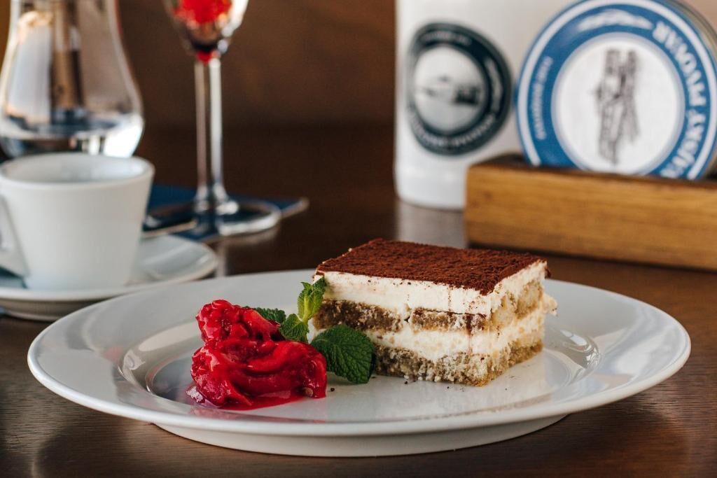 Uma fatia de tiramisu em um prato branco, com uma rosa vermelha e folhas de hortelã.