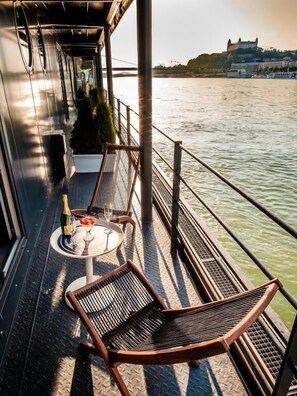 A deck with two wicker chairs, a small round table with a bottle and glass, and a view of a castle across the water. - Botel Dunajský Pivovar (Bratislava)