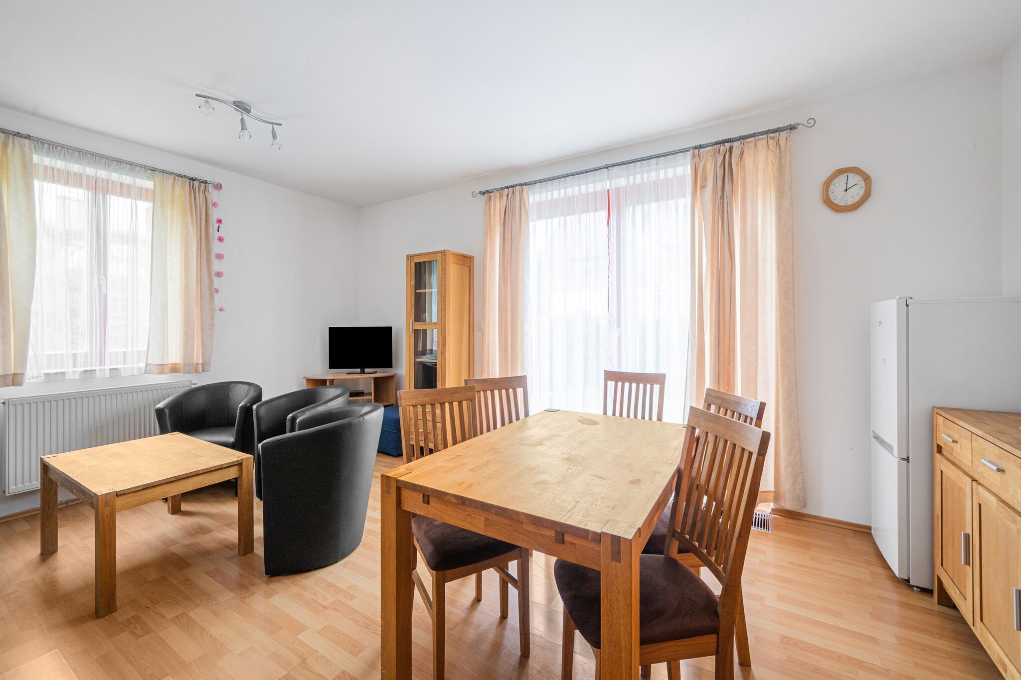 A spacious living area with a wooden dining table and chairs, a sofa, a television, and a kitchen area with a refrigerator.
