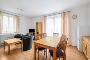 A spacious living area with a wooden dining table and chairs, a sofa, a television, and a kitchen area with a refrigerator.