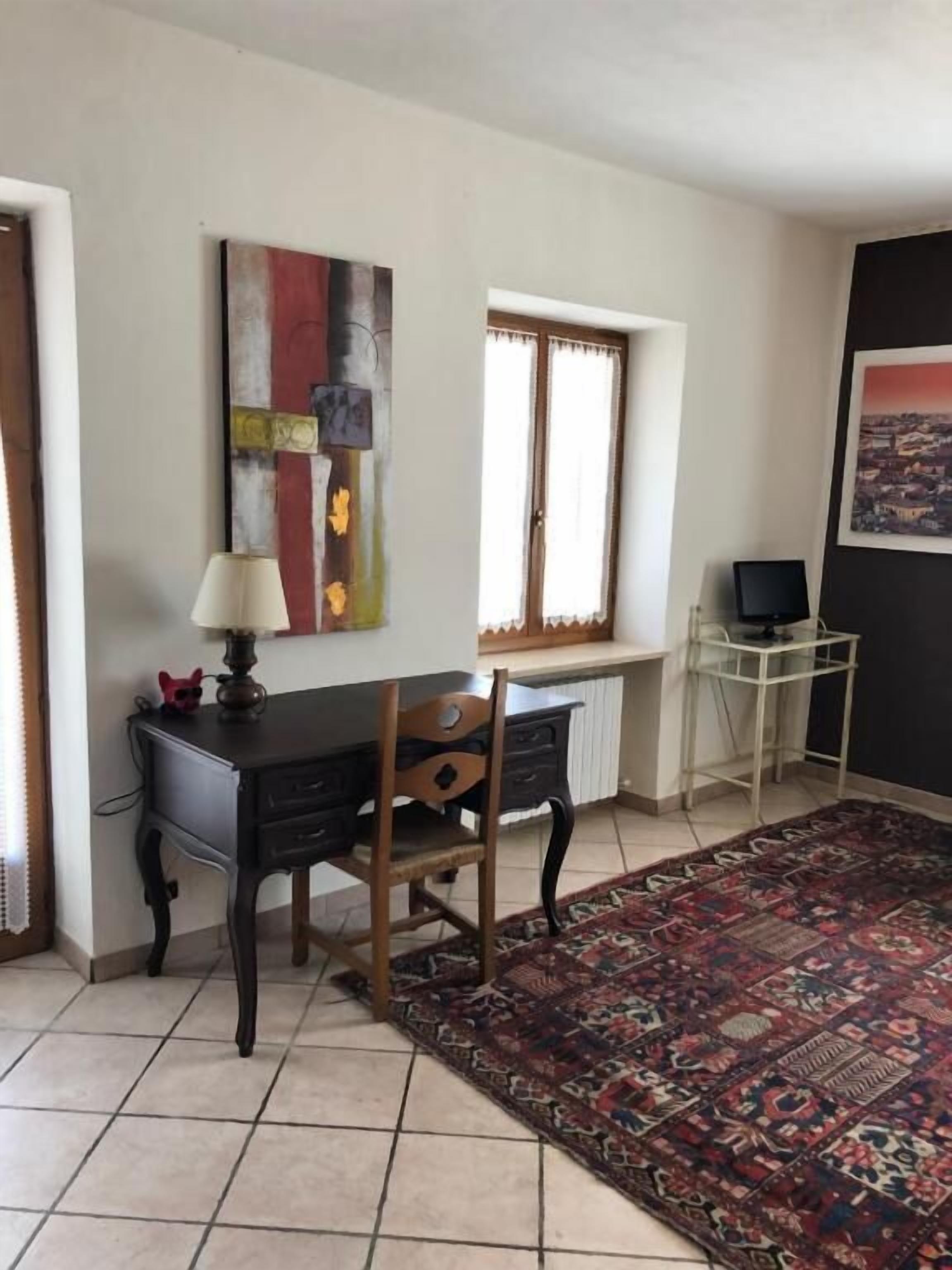 A room with a desk, chair, and a large rug. There is a window with curtains, a radiator, and a framed picture on the wall.