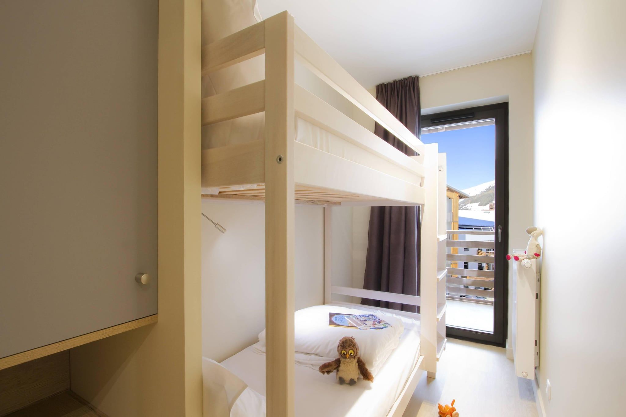 A hotel room with a bunk bed, a small table with a teddy bear, and a window with a view of a mountain.