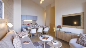 Een moderne woonkamer met een bank, een salontafel, een aan de muur bevestigde televisie en een eethoek met stoelen en een tafel.