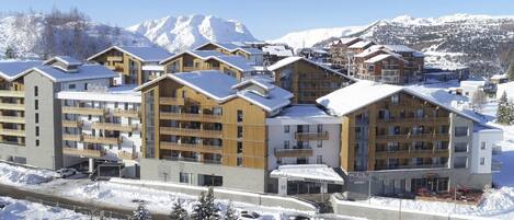 A snow-covered mountain resort with multi-story buildings, ski lifts, and a clear view of the surrounding mountains.