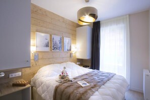 A hotel room with a large bed, a wooden headboard, wall-mounted artwork, a bedside table with a lamp, and a window with curtains. - Apartment in France with Mountain Views (Huez)