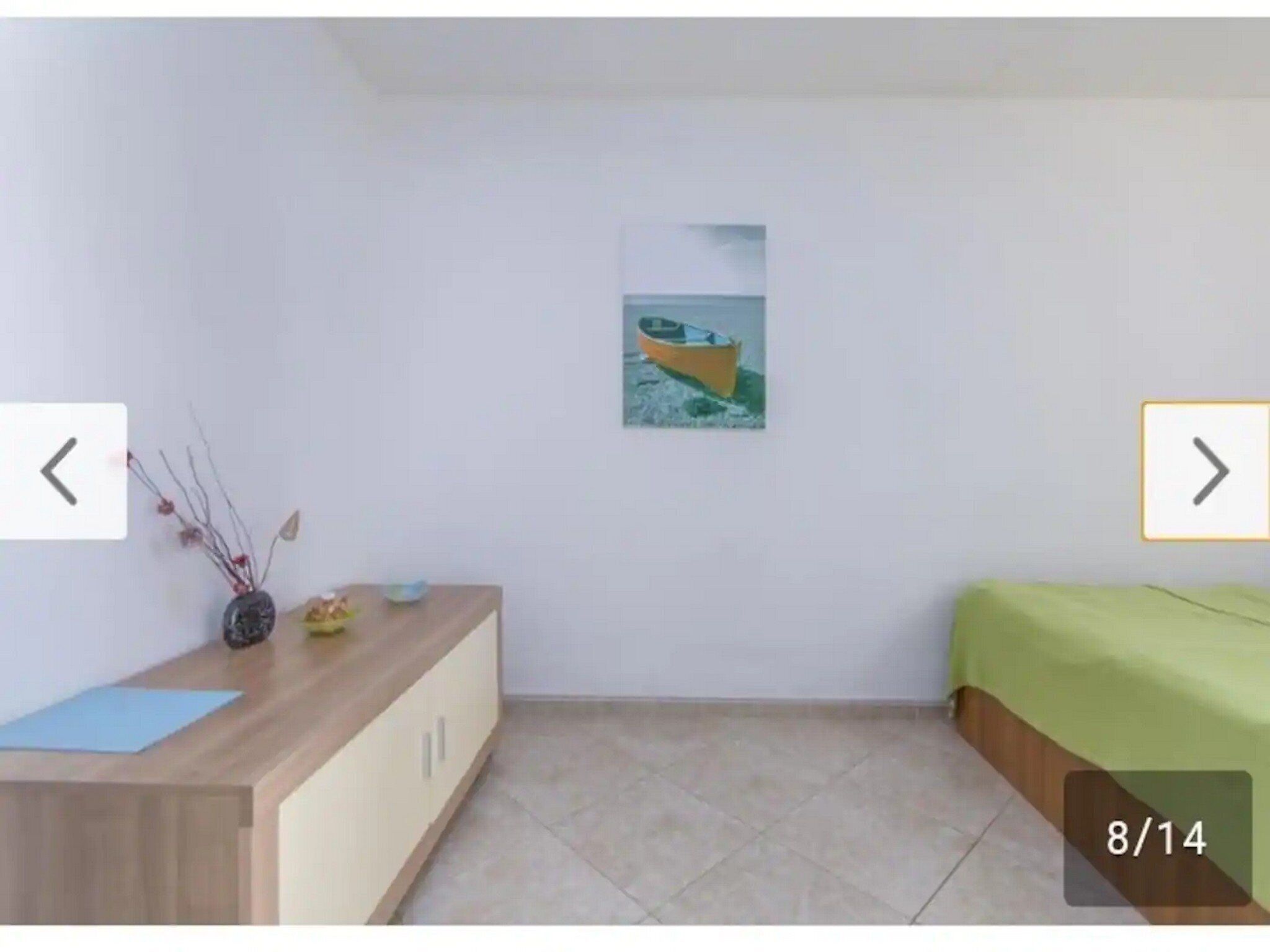 A small, clean room with a wooden dresser, a bed with green bedding, and a painting of a boat on the wall.