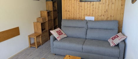 A cozy room with a grey sofa, wooden shelves, a small table, and a heart-shaped chair.