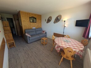 Een gezellige woonkamer met een bank, een eettafel met geruit tafelkleed en een aan de muur bevestigde televisie.