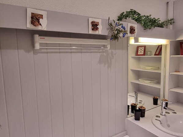 Une salle de bain avec un meuble-lavabo blanc, un évier et un miroir. Des tableaux encadrés ornent le mur et une plante est placée au-dessus du miroir.