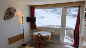 Een knus ingerichte kamer met een ronde tafel, houten stoelen, een klein houten krukje, een flatscreen televisie en een groot raam met uitzicht op een besneeerd landschap.