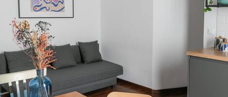 A modern living room with a wooden dining table, grey sofa, and a kitchen area with a shelf.