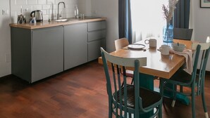 Una cuina compacta amb taulell de fusta, armaris grisos i una prestatgeria integrada. Hi ha una taula de dinar amb caires, un vas amb flors i una finestra amb cortines.