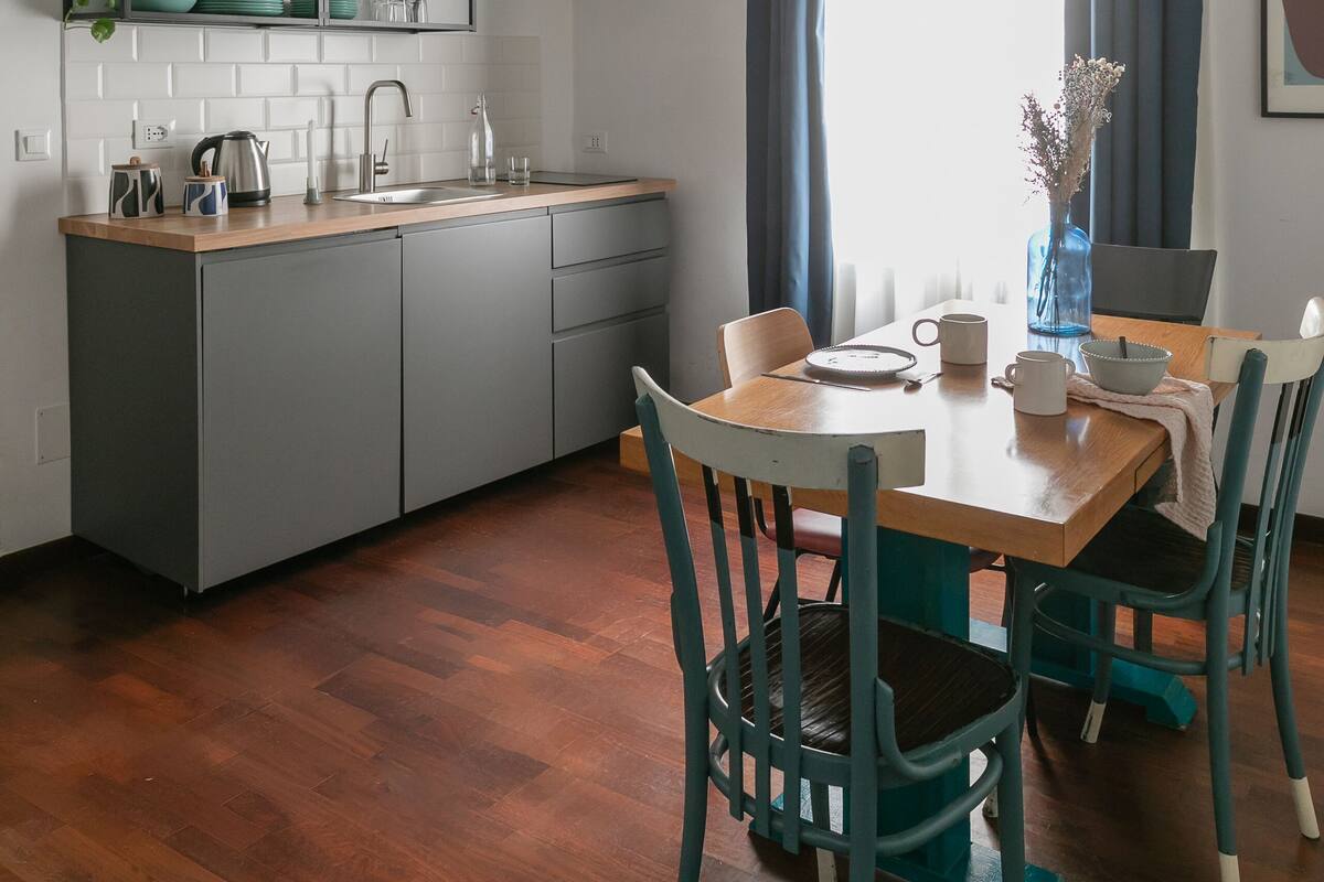 A compact kitchen with a wooden countertop, grey cabinets, and a built-in shelf. There is a dining table with chairs, a vase with flowers, and a window with curtains.