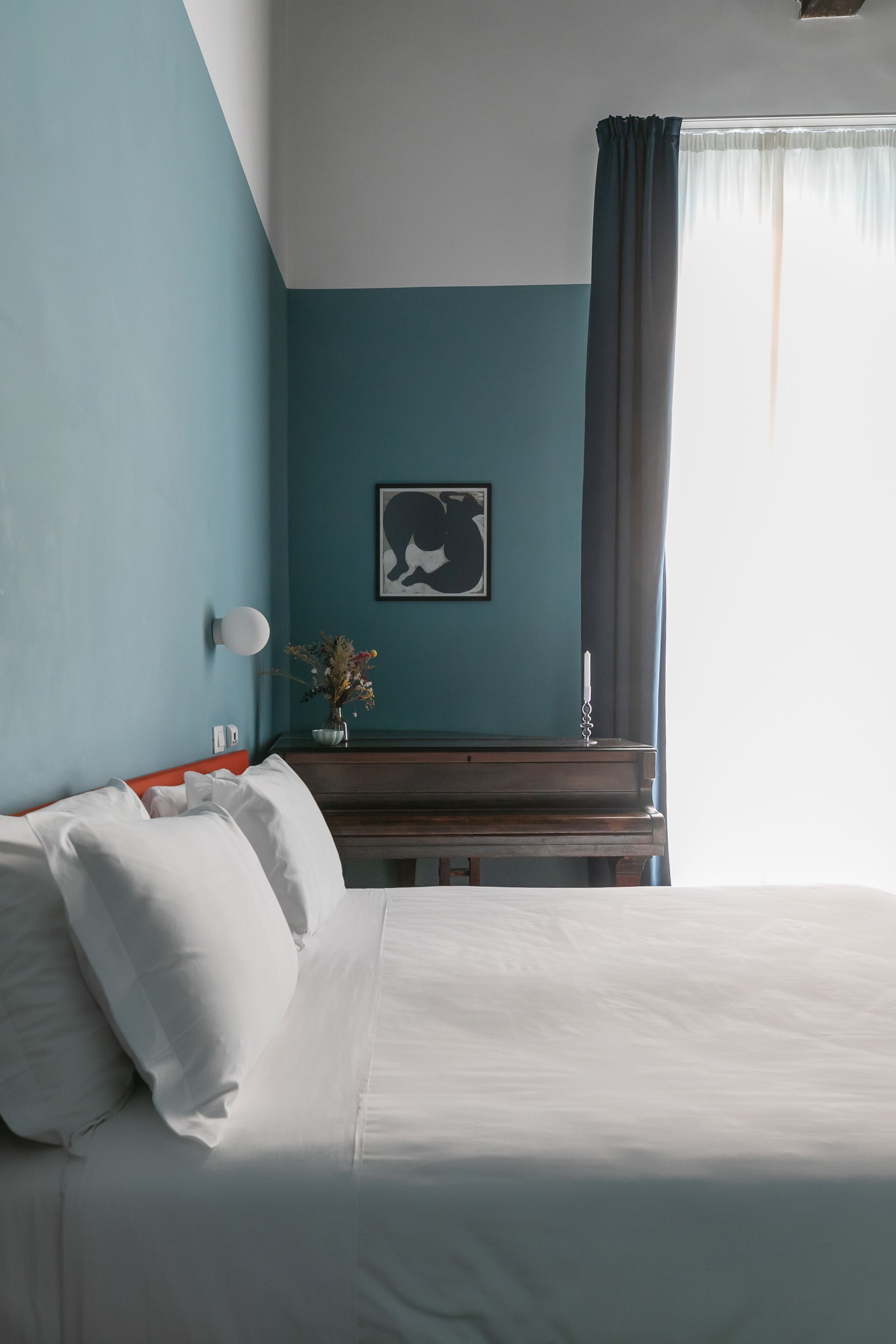 Hotelkamer met een bed, nachtkastje, een wandlamp, een lijst met een afbeelding en een raam met gordijnen.