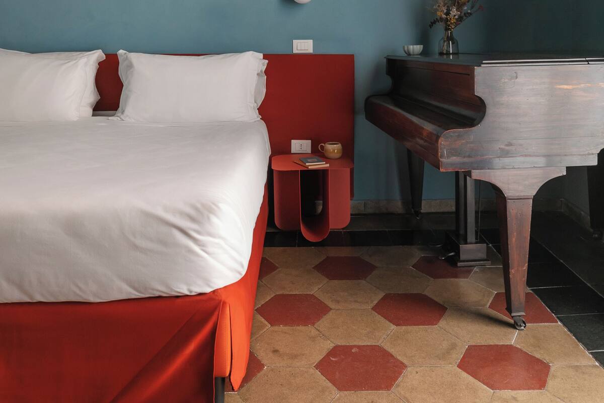 A hotel room with a bed, a red nightstand, a piano, and a vase with flowers.