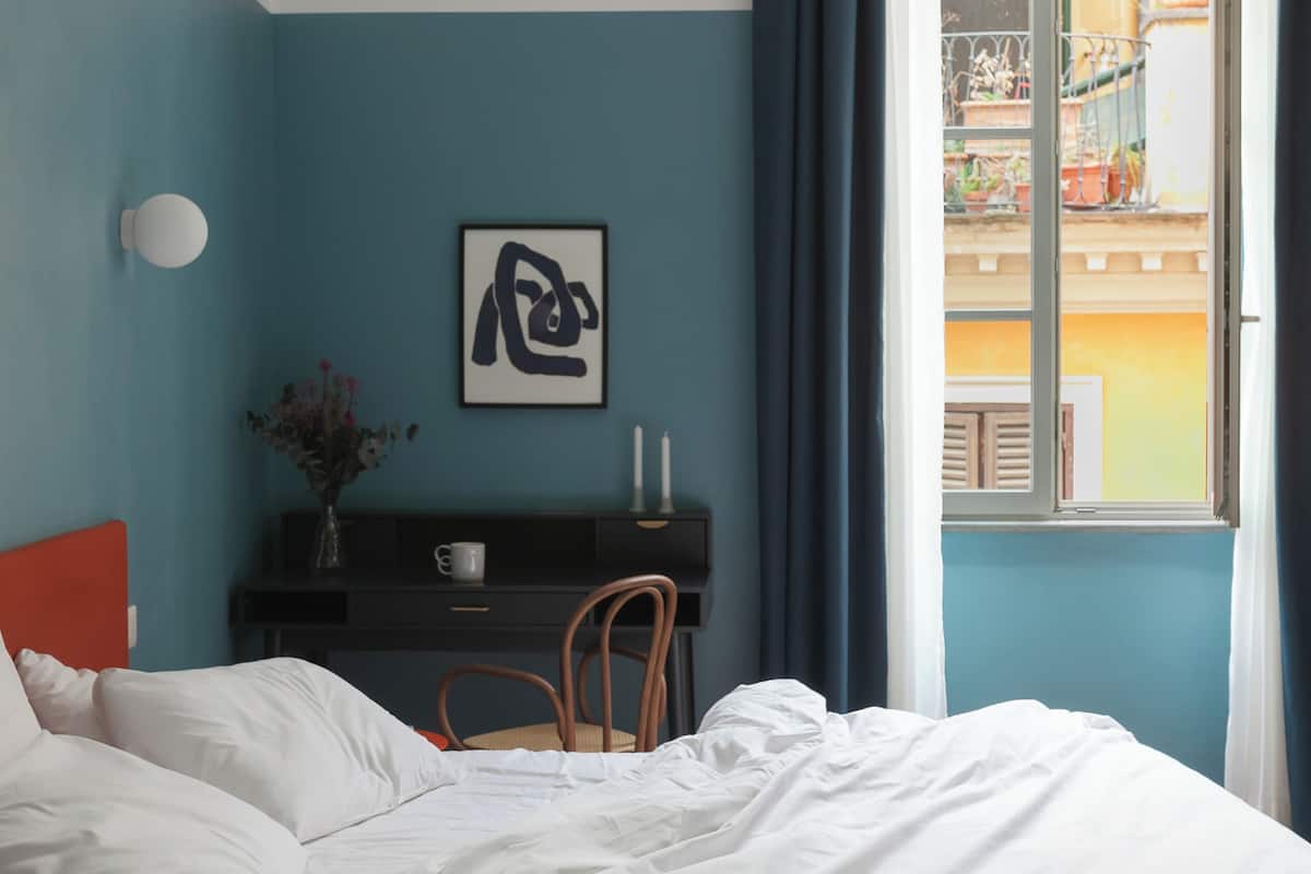 A bedroom with a bed, a desk, a chair, a vase with flowers, and a window with curtains.