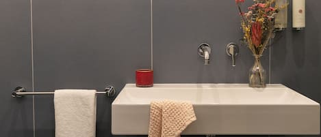 Un baño con un lavabo blanco, baldosas oscuras, un perchero, un florero con flores y un espejo.