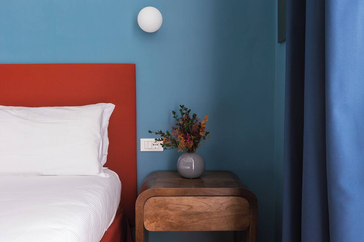 A neatly made bed with white bedding, a small wooden nightstand with a vase of flowers, and a wall socket.