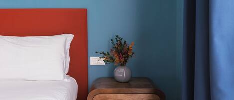 A neatly made bed with white bedding, a small wooden nightstand with a vase of flowers, and a wall socket.