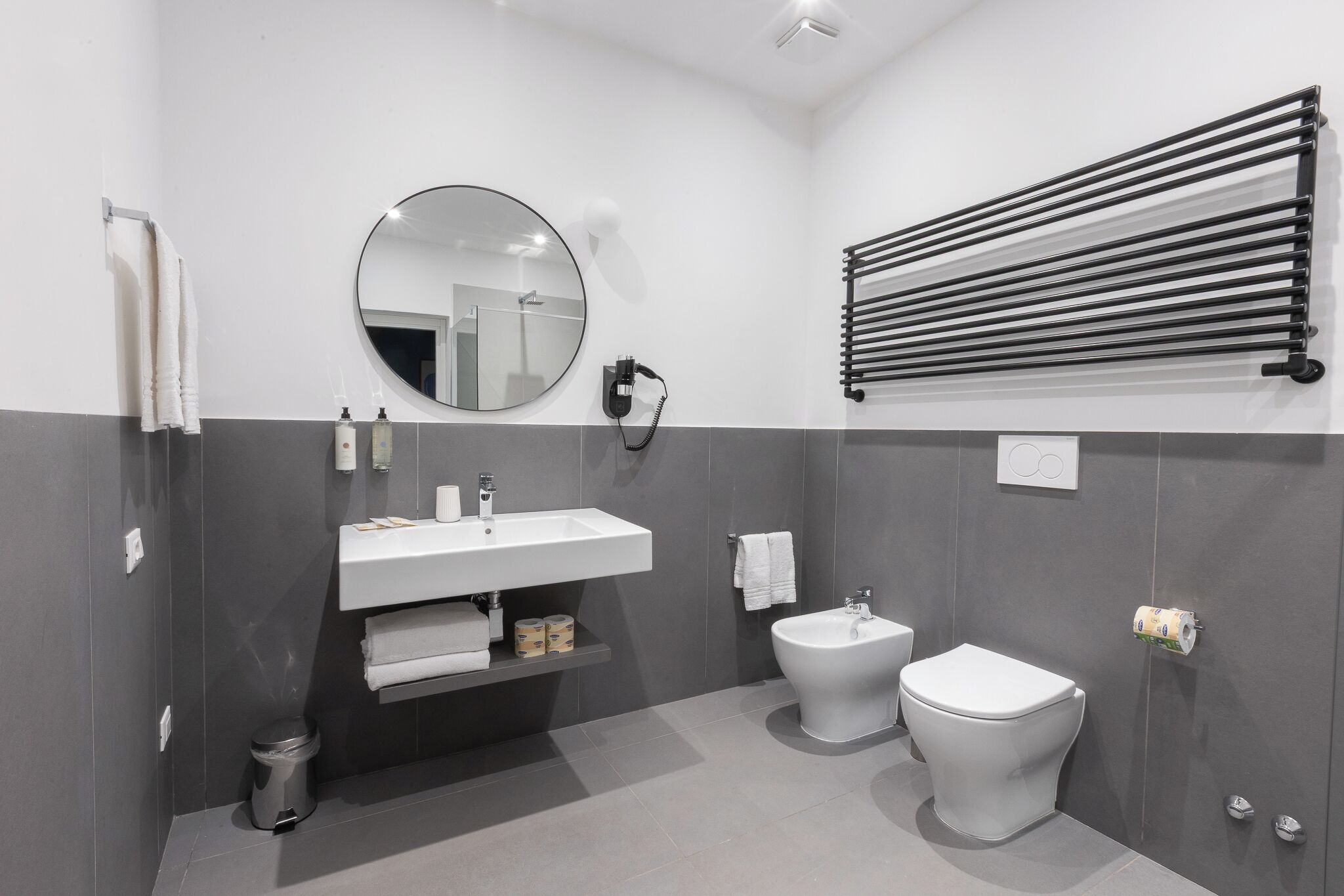 A modern bathroom with a white sink, a round mirror, a bidet, and a towel rack.