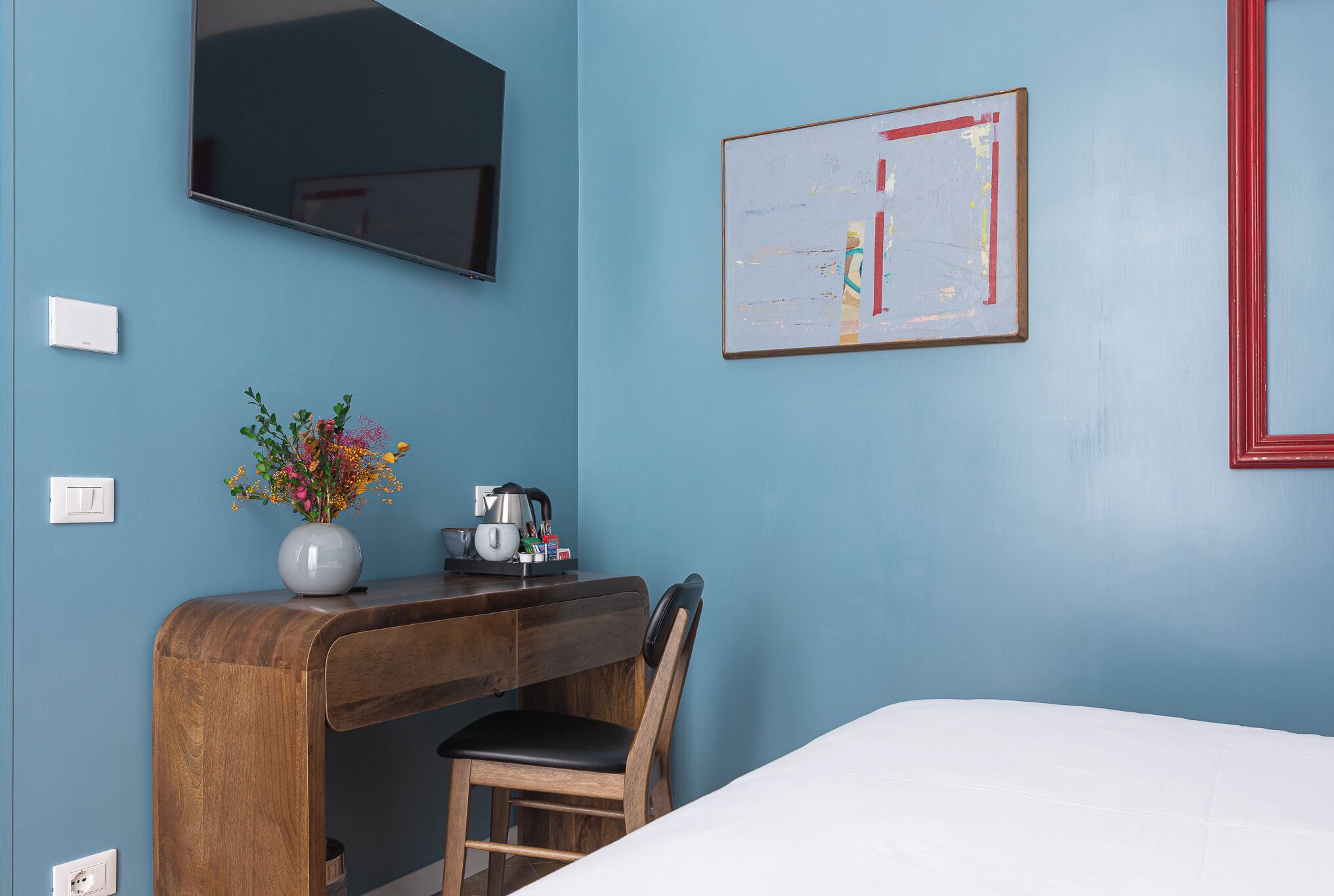 A hotel room with a wooden desk, a chair, a bed, a TV mounted on the wall, and a framed picture.