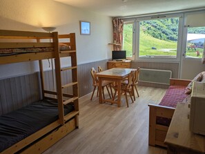 A bunk bed, a wooden table with chairs, a television, and a window with a view of greenery.