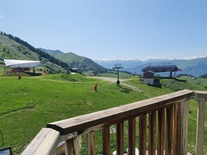 A scenic view of a mountainous area with a wooden deck overlooking a green field, a white structure, and a red building in the distance.