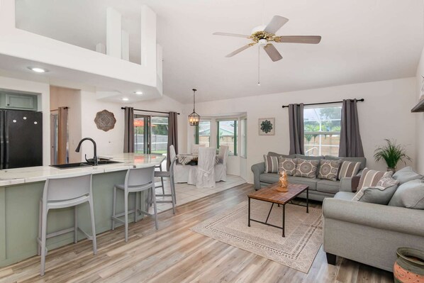 A modern kitchen with a central island, a dining area with chairs, a sofa, and a ceiling fan.