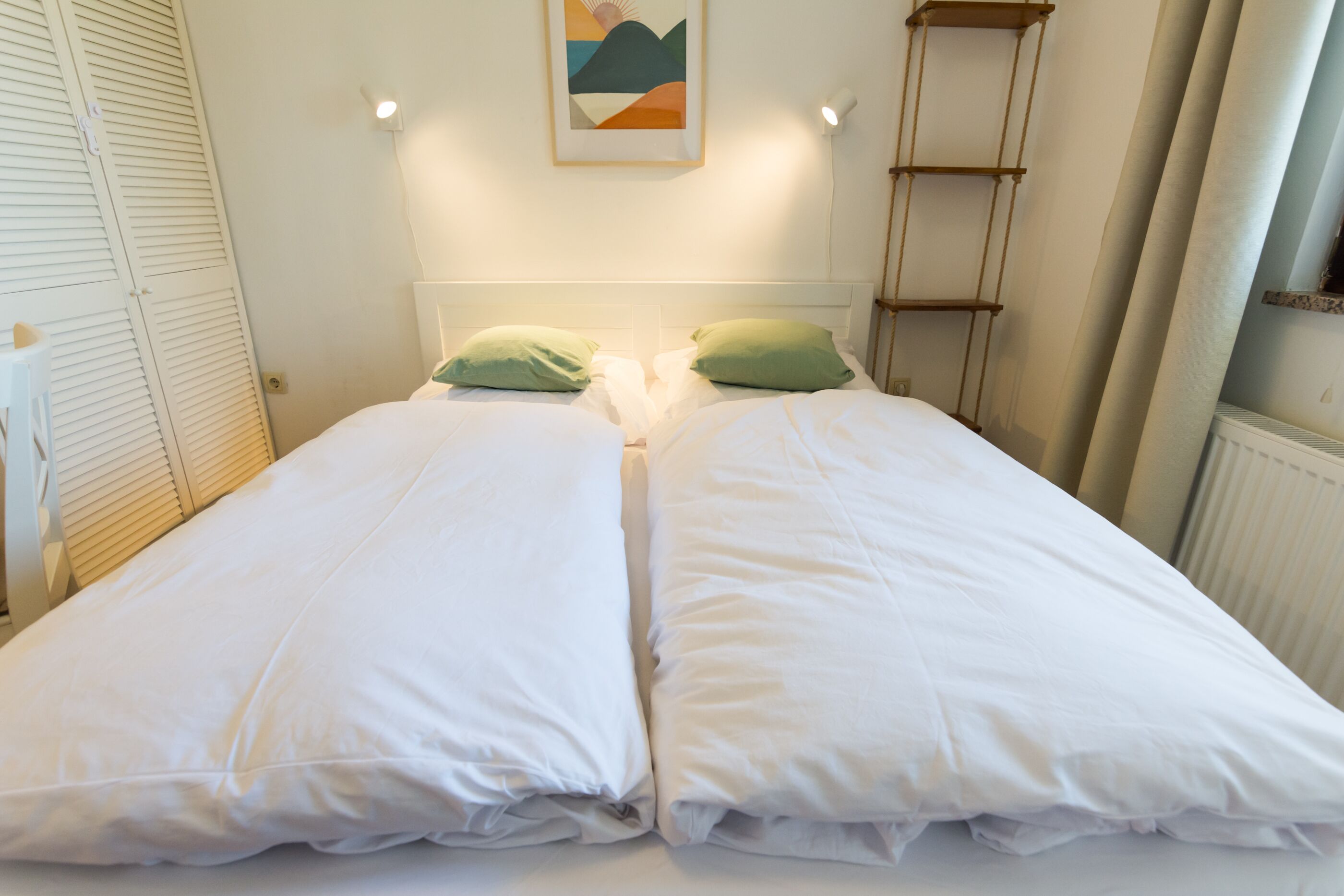 Un lit double avec une literie blanche et des oreillers verts, une œuvre d’art encadrée au-dessus du lit, et une échelle appuyée contre le mur.