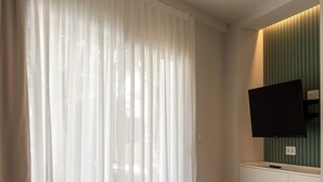 A hotel room with a large window covered by sheer curtains, a flat-screen TV mounted on a green wall, and a checkered floor pattern. - SUNWAY ATLANTA (Sitges)