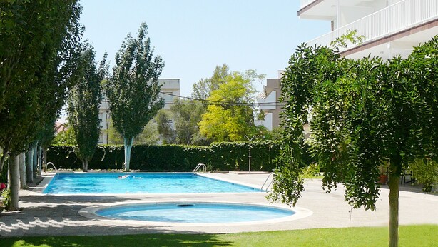 A swimming pool surrounded by well-maintained grass and trees, with a building featuring balconies in the background. - SUNWAY ATLANTA (Sitges)