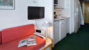 A compact kitchen with a microwave, stove, and sink. A red sofa with a magazine is in the foreground.